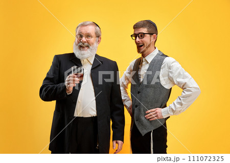 Two happy and joy, young and mature men posing in traditional costumes holds glass of alcohol drink against yellow background. Happy Purim. 111072325