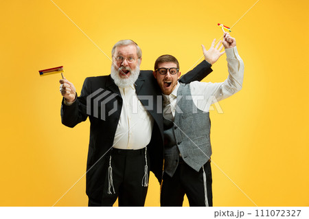 Two Jewish men, mature and young celebrating start of celebrating of national holiday against vivid yellow background. Happy Purim. Two Jewish men, mature and young celebrating start of celebrating of national holiday against vivid yellow background. Happy Purim. 111072327