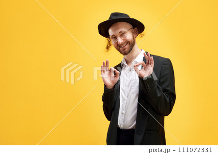 Portrait of smiling man in suit and black hat making positive hand gesture against sunny yellow background. National holidays. Portrait of smiling man in suit and black hat making positive hand gesture against sunny yellow background. National holidays. 111072331