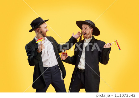 Two Jewish men, friends happy to meet at Purim holiday celebration. Male model posing with grogger and alcohol drinks against yellow background. Two Jewish men, friends happy to meet at Purim holiday celebration. Male model posing with grogger and alcohol drinks against yellow background. 111072342