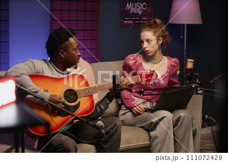 African American male musician playing acoustic guitar sitting on couch with young Caucasian female podcaster holding laptop in studio with neon light African American male musician playing acoustic guitar sitting on couch with young Caucasian female podcaster holding laptop in studio with neon light 111072429