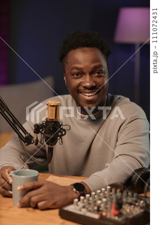 Portrait of happy Black man looking at camera holding cup sitting at table with audio equipment in podcast studio 111072431
