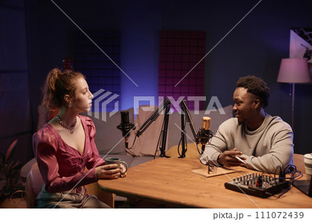 Smiling African American host at desk with studio equipment interviewing fashionable Caucasian woman holding cup in comfortable neon lit podcast room Smiling African American host at desk with studio equipment interviewing fashionable Caucasian woman holding cup in comfortable neon lit podcast room 111072439