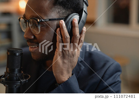 Close up view of smiling African American man wearing glasses adjusting headphones while recording podcast in studio Close up view of smiling African American man wearing glasses adjusting headphones while recording podcast in studio 111072481