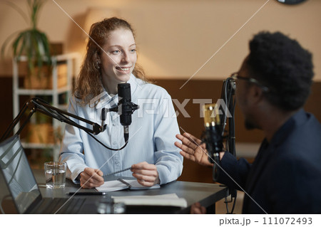 Medium shot of cheerful Caucasian girl listening to African American male podcast guest while sitting at table in studio 111072493
