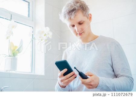 Young man puts the settings of his electronic toothbrush with mobile phone app. Wireless connecting sonic toothbrush with smart phone app. Modern home health care technology concept. Selective focus Young man puts the settings of his electronic toothbrush with mobile phone app. Wireless connecting sonic toothbrush with smart phone app. Modern home health care technology concept. Selective focus 111072679