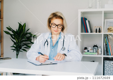 Confident mature middle aged woman doctor medical nurse, female physician practitioner in white coat with stethoscope sitting at working place at hospital office and making notes during appointment. 111072689
