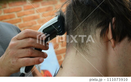 a man's hand holds a hair clipper with a nozzle and cuts the back of a young man's head, close-up, men in the family take care of each other, home haircut of a teenager using a trimmer a man's hand holds a hair clipper with a nozzle and cuts the back of a young man's head, close-up, men in the family take care of each other, home haircut of a teenager using a trimmer 111073910