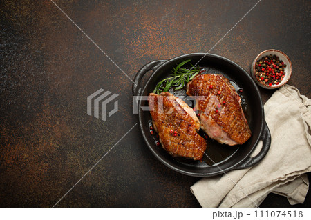 Two roasted duck breast fillets with crispy skin, with pepper and rosemary, top view in black cast iron pan with knife, dark brown concrete rustic background, space for text 111074518