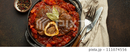 Traditional Italian dish Ossobuco all Milanese made with cut veal shank meat with vegetable tomato sauce served in black casserole pan top view on rustic brown background 111074556