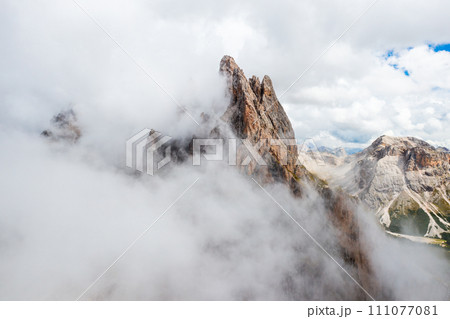 Dense fog descends onto Seceda mountain peaks on sunny summer day. Giant ridgeline with bare peak and steep slope in Italian Alps aerial view 111077081