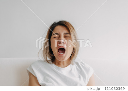 Sleepy face woman yawning and rubbing her eyes in white t-shirt sits on the sofa Sleepy face woman yawning and rubbing her eyes in white t-shirt sits on the sofa 111080369