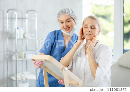 Happy elderly woman examining face after procedures in cosmetology clinic Happy elderly woman examining face after procedures in cosmetology clinic 111081875
