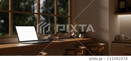 A white-screen laptop mockup on a hardwood desk against the window in a modern, cozy kitchen. A white-screen laptop mockup on a hardwood desk against the window in a modern, cozy kitchen. 111082074