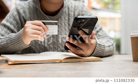 A cropped shot of a woman holding her smartphone and a credit card while sitting at a table. 111082330