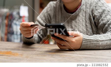 A cropped shot of a woman holding her smartphone and a credit card while sitting at a table. 111082332