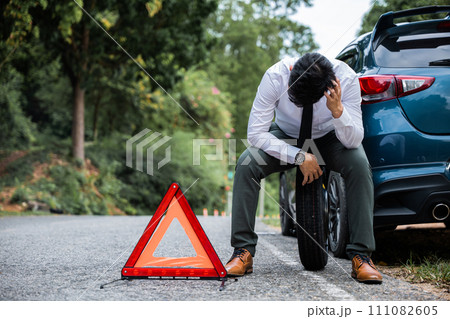 A young businessman in a white shirt confidently changing the wheel of a broken car while a worried mature man sits beside him. A red triangle sign warns drivers of the obstacle ahead. A young businessman in a white shirt confidently changing the wheel of a broken car while a worried mature man sits beside him. A red triangle sign warns drivers of the obstacle ahead. 111082605