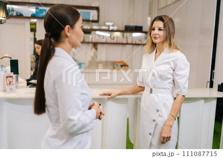 Side view of two female doctors standing on hospital corridor discussing patient diagnose. Young physician consulting woman colleague standing by reception. Concept of healthcare. 111087715