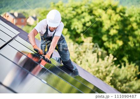 Worker building photovoltaic solar panel system on rooftop of house. Man engineer in helmets and gloves installing solar module with help of hex key outdoors. Alternative energy generation concept. Worker building photovoltaic solar panel system on rooftop of house. Man engineer in helmets and gloves installing solar module with help of hex key outdoors. Alternative energy generation concept. 111090487