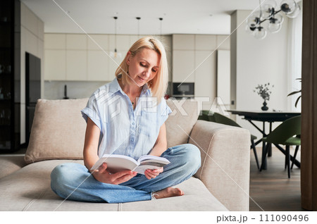 Woman sitting comfortably on couch, deeply engaged in reading book. Cozy living space with modern aesthetic on background. Person enjoying quiet moment of solitude, immersed in reading. 111090496