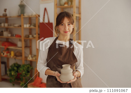 Portrait of young female potter in apron with mug looking at camera while posing in workshop Portrait of young female potter in apron with mug looking at camera while posing in workshop 111091067