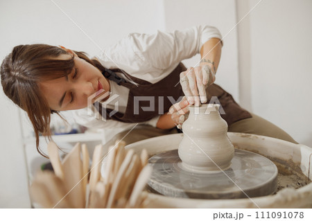 Smiling female artisan in apron sitting on bench with pottery wheel and making clay pot 111091078
