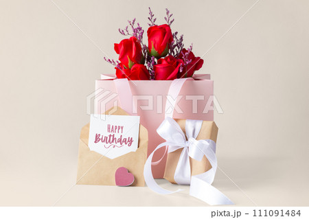 Happy birthday card and envelope with gift box and rose in paper bag on beige background Happy birthday card and envelope with gift box and rose in paper bag on beige background 111091484