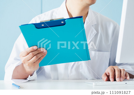 ファイルとパソコン画面を見比べる男性医師の手元 111091827