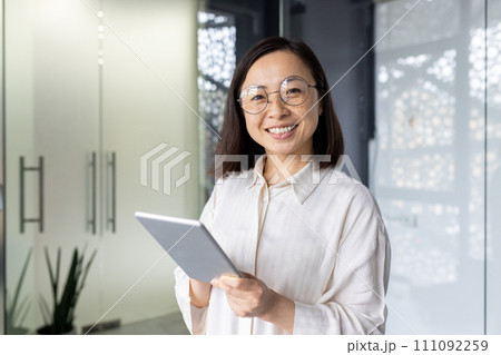 Professional young Asian businesswoman with a tablet, smiling confidently at her workplace desk in a contemporary office setting. 111092259