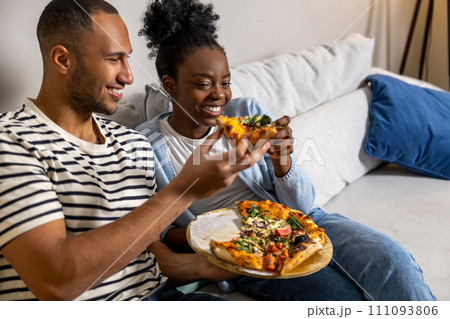 Couple eating pizza snack at home and laughing while resting on sofa Couple eating pizza snack at home and laughing while resting on sofa 111093806