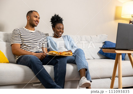 Woman and man enjoying pizza for dinner watching movie on computer sitting on couch at home 111093835