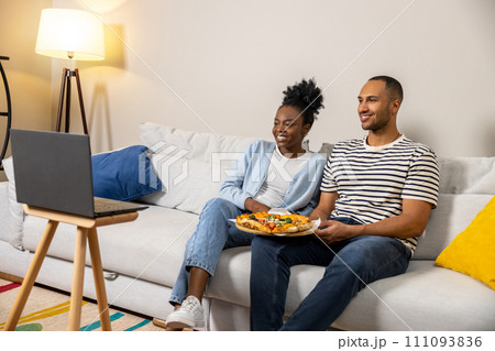 Male and female couple eating pizza while watching movie on laptop and sitting on couch 111093836