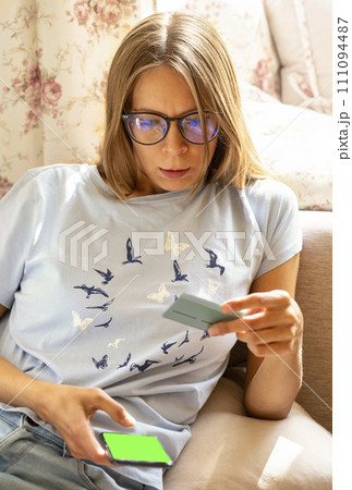 Young woman sitting at home with smartphone with green screen and bank card in hands. Online payment 111094487