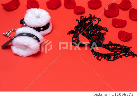 Valentine's day black lace mask and fur handcuffs on red background with rose petals Valentine's day black lace mask and fur handcuffs on red background with rose petals 111096573