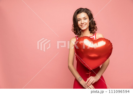 Portrait of young lady stands on pink background with heart shapped red balloon Portrait of young lady stands on pink background with heart shapped red balloon 111096594