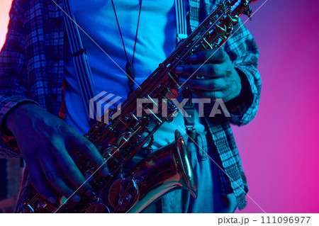 Cropped portrait of jazzman holds saxophone against gradient blue-pink background in neon light. Jazz melodies. Cropped portrait of jazzman holds saxophone against gradient blue-pink background in neon light. Jazz melodies. 111096977