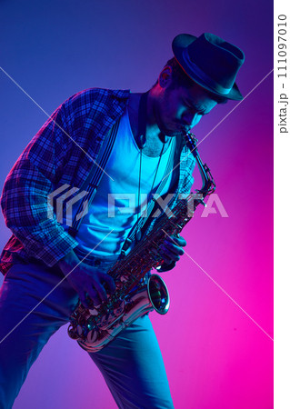 African-American man dressed retro, in hat playing saxophone against gradient pink and blue background in neon light. 111097010
