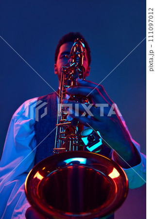 Close up portrait. African-American talented man, musician playing saxophone against studio background in blue and pink lighting. 111097012