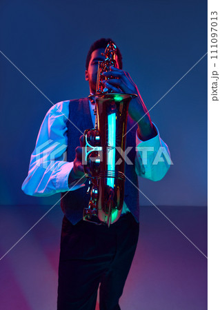 Jazzman. Elegant saxophonist dressed white shirt and vest playing jazz melodies in blue-pink light against gradient studio background. 111097013