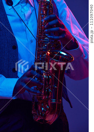 Cropped photo of African-American jazz-man playing saxophone, wind instrument against gradient background in mixed neon light. Cropped photo of African-American jazz-man playing saxophone, wind instrument against gradient background in mixed neon light. 111097040