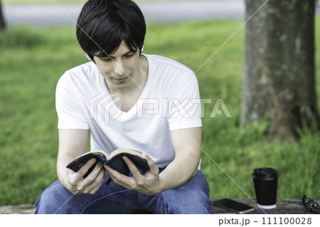 新緑の公園で音楽を聴きながら読書をする外国人の男性 111100028
