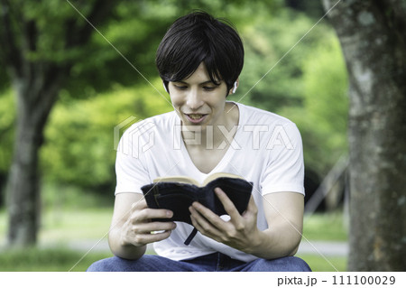 新緑の公園で音楽を聴きながら読書をする外国人の男性 111100029