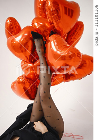 Cropped photo of legs of woman with heart shape air balloons on a white background. Woman on Valentine's Day wearing a black tights and dress. Woman in studio posing for a photo. 111101106