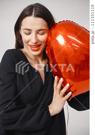 A beautiful young caucasian woman with heart shape air balloon on a white background. Woman on Valentine's Day wearing a black dress. Brunette woman in studio posing for a photo. A beautiful young caucasian woman with heart shape air balloon on a white background. Woman on Valentine's Day wearing a black dress. Brunette woman in studio posing for a photo. 111101119