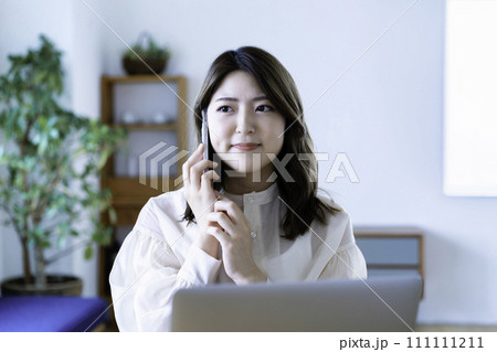 リビングでノートパソコンを使いながらスマホ通話する若い女性 111111211