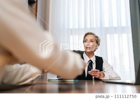 View from desk of middle-aged businesswoman manager passing papers documents to unrecognizable female colleague, sitting at table in office. Two business partners having meeting in conference room. 111112750