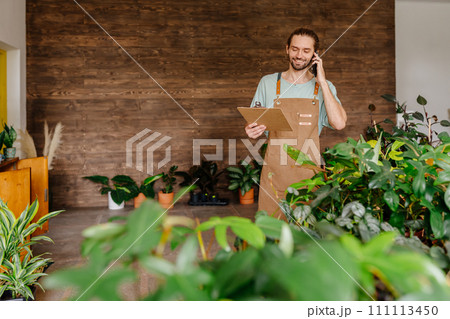 Smiling seller talking on smartphone and writing notes on clipboard in flower shop. Floristry business and people concept. Smiling seller talking on smartphone and writing notes on clipboard in flower shop. Floristry business and people concept. 111113450