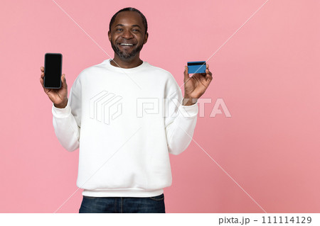 Cheerful black man wearing white sweatshirt showing phone with blank screen and credit card Cheerful black man wearing white sweatshirt showing phone with blank screen and credit card 111114129