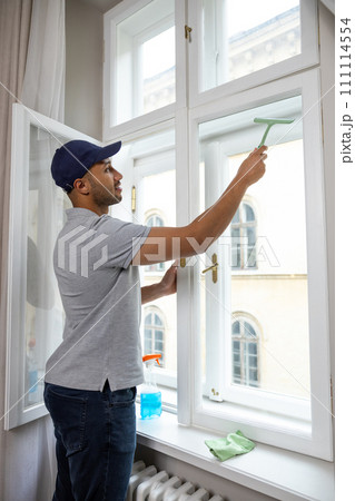Young man in cap and T-shirt washing window at home with wiper squeegee. 111114554
