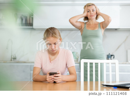 Teen girl fixated on phone while dissatisfied mother standing in background 111121408